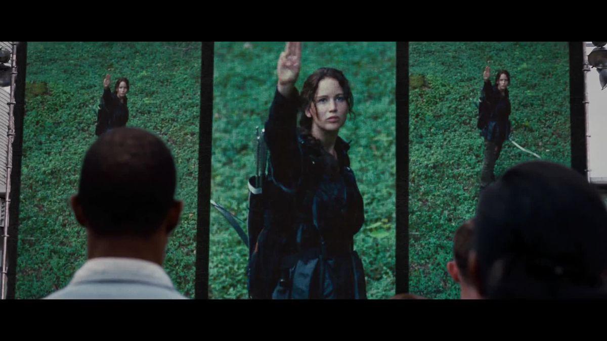 Several people seen from behind watch on a series of monitors as Katniss Everdeen gives the Mockingjay hand sign in The Hunger Games