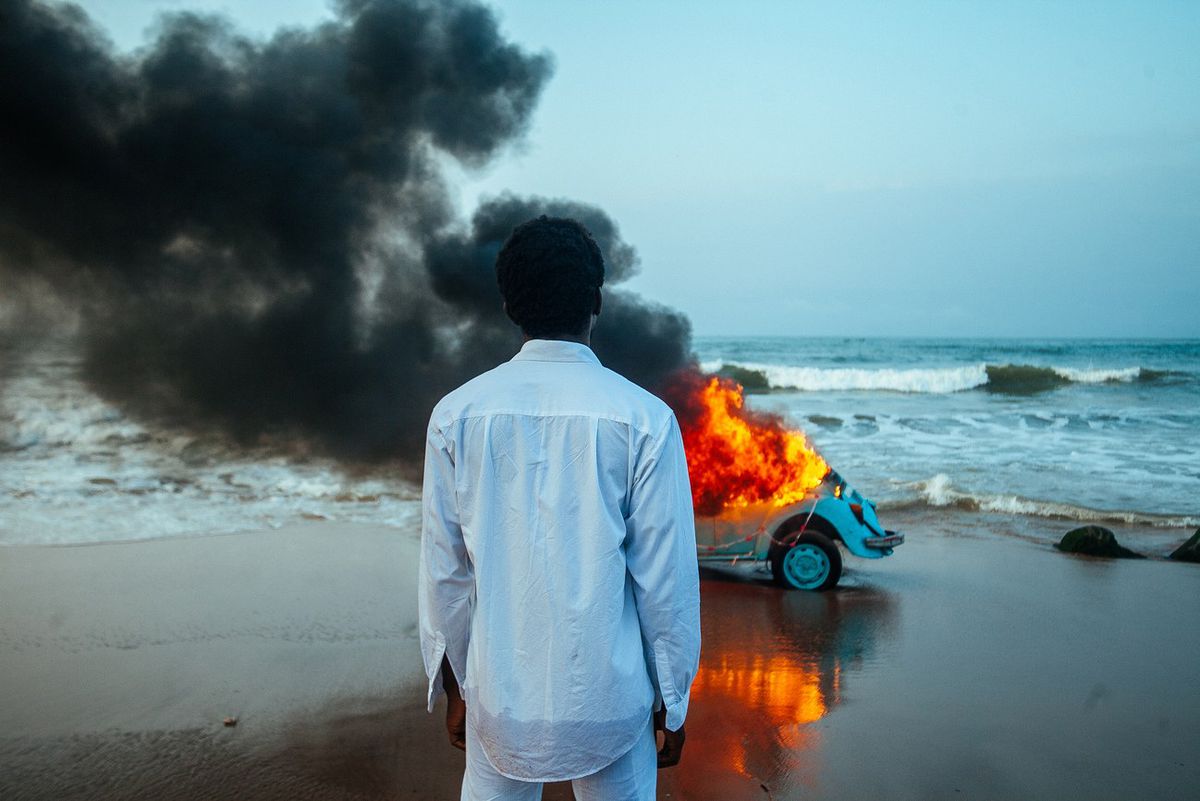The Burial of Kojo: A Ghanian man stands in front of a burning Volkswagen beetle