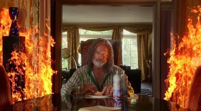Kelsey Grammer sits at the far end of a kitchen table eating a sandwich as roaring flames rage around him.