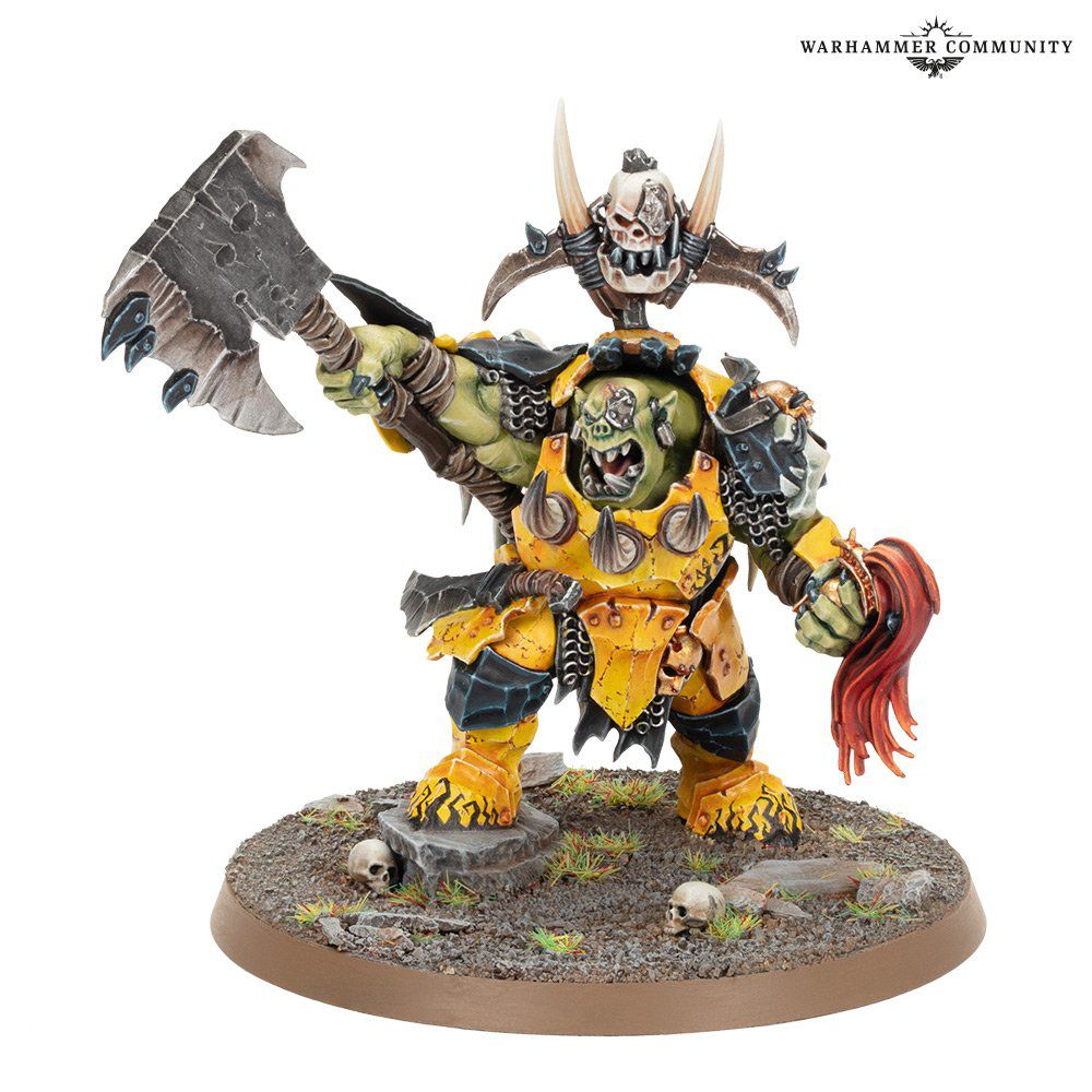 Warhammer Fantasy - a growling orc lifts his axe and menaces enemies while holding the helmet of one of Sigmar’s warriors.