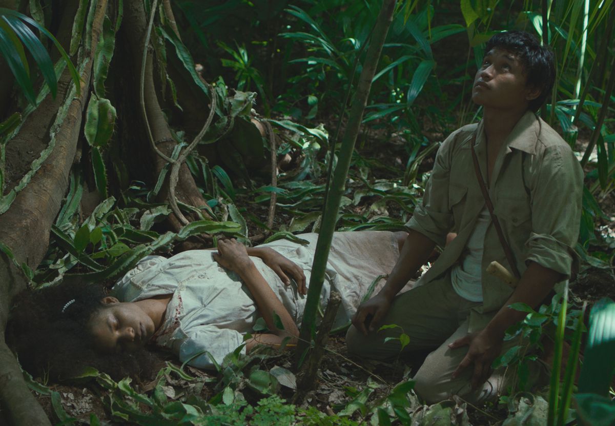 A woman in white lies in the jungle as a young man kneels next to her, looking up
