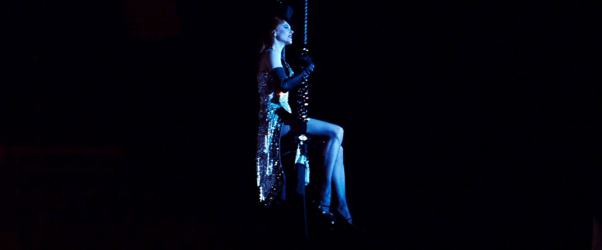 Nicole Kidman, in a spangled leotard, cape, and tophat, sits on a swing in a dark room in Moulin Rouge