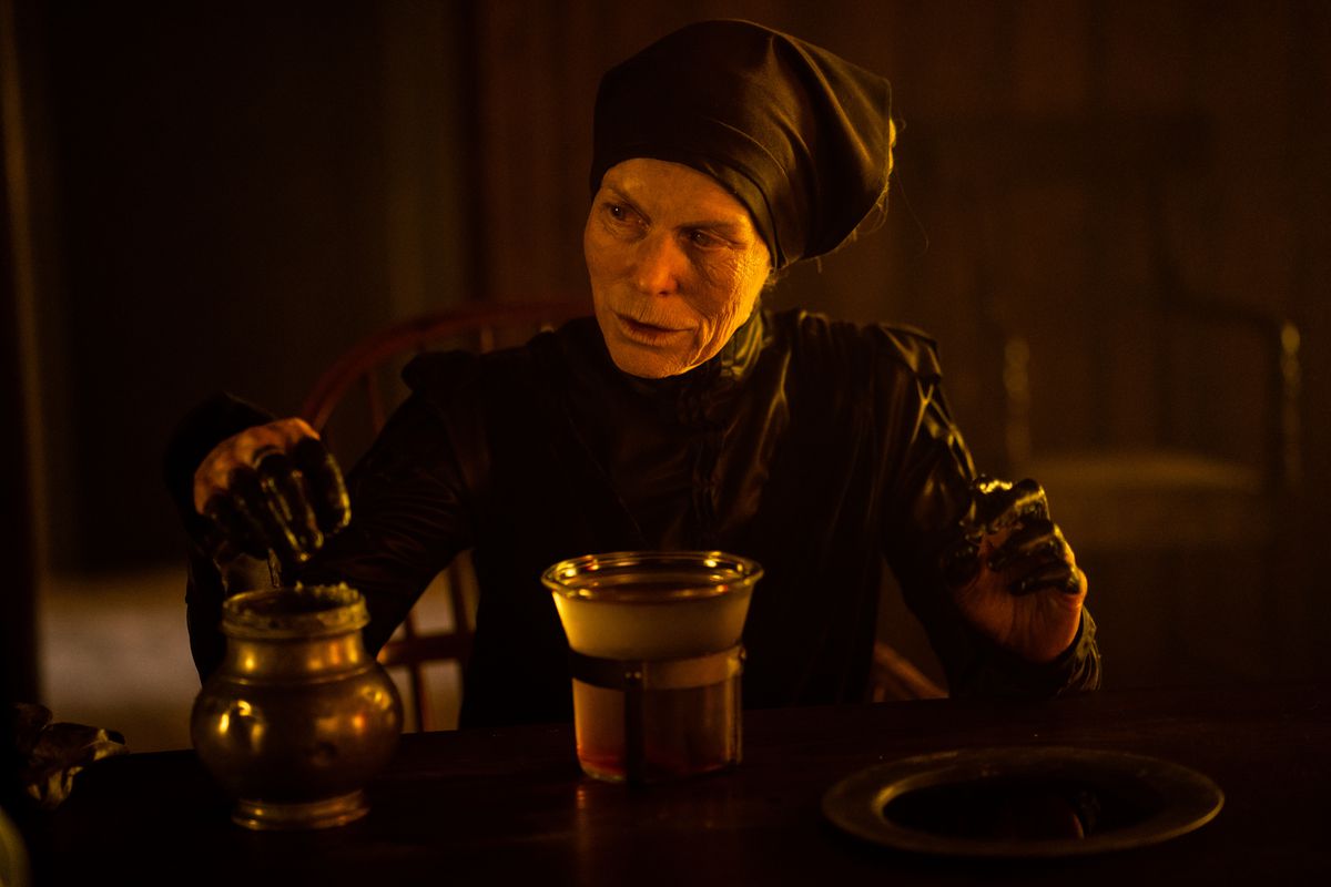 The Witch in Gretel and Hansel, an elderly woman dressed in a black robe and head-wrap, dips her blackened fingers into a brass pot full of goo.