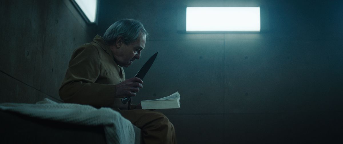 An elderly man clutching a nasty-looking knife and reading a book sits on his bed in a dimly lit concrete cell in The Platform.