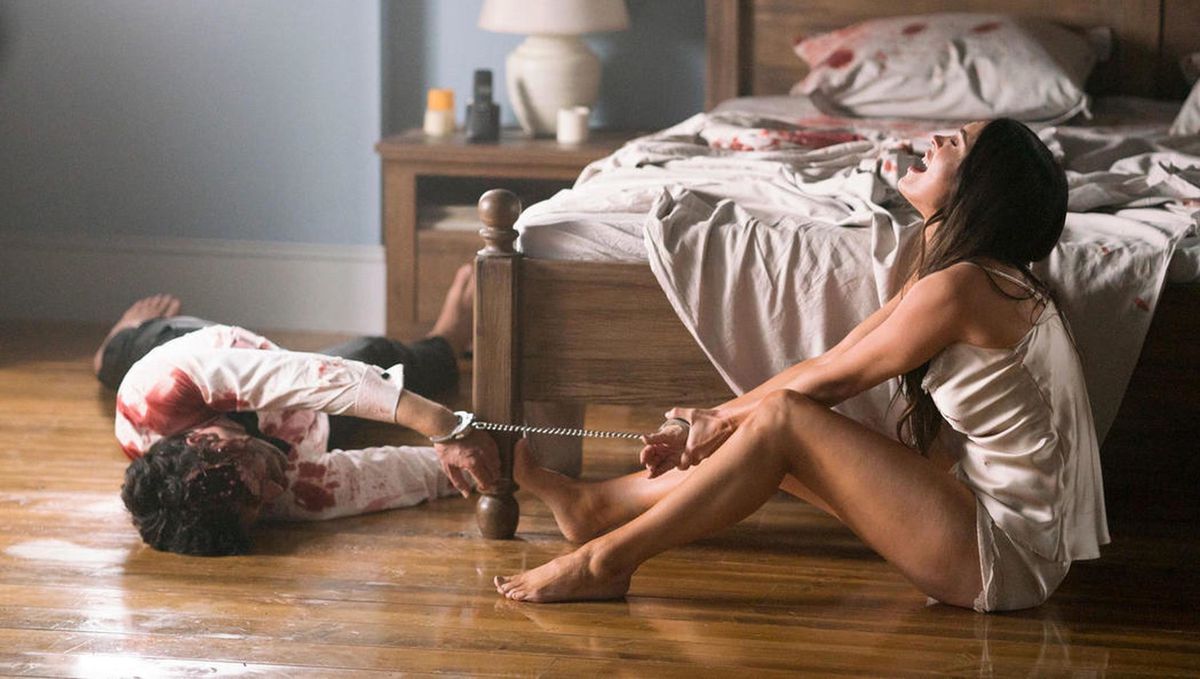 Megan Fox, in a silk camisole and underwear, weeps on the floor at the foot of a bed while handcuffed to the corpse of a bloody man in Till Death.