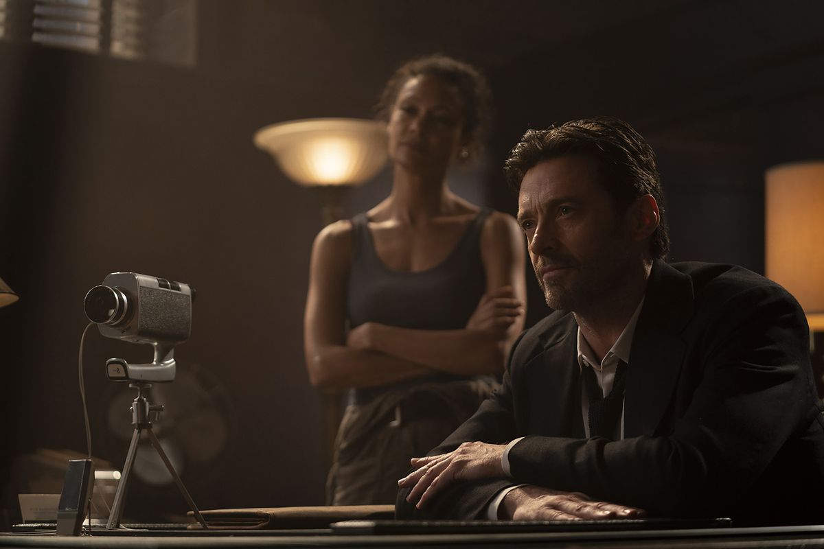 Hugh Jackman sits at a table with a video camera pointed at his offscreen subject while Thandiwe Newton stands behind him in Reminiscence