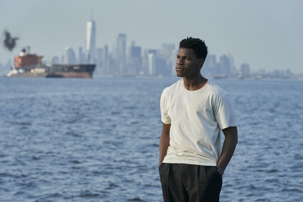 John Boyega, slouching in a white T-shirt and loose black pants, stands by water with Manhattan in the background in Naked Singularity