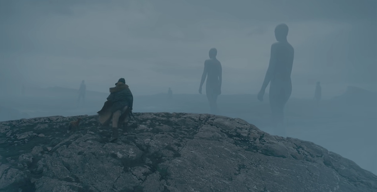 Gawain (Dev Patel) stands at the edge of a cliff, looking down into a misty grey valley where vast naked giants are walking toward the horizon